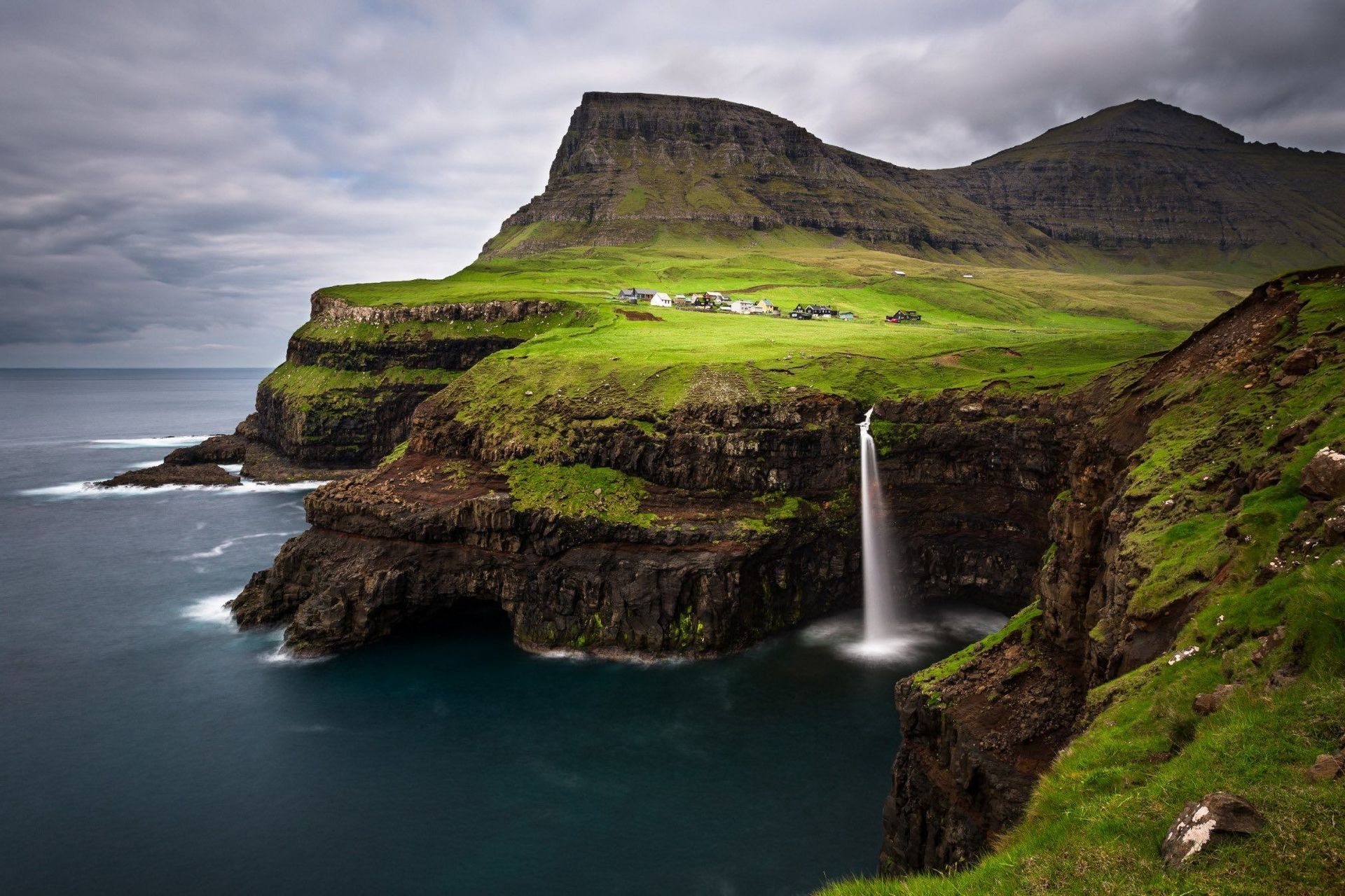 Una cascata si getta da una rigogliosa scogliera verde direttamente nell'oceano, con un piccolo villaggio annidato sulla collina sullo sfondo.