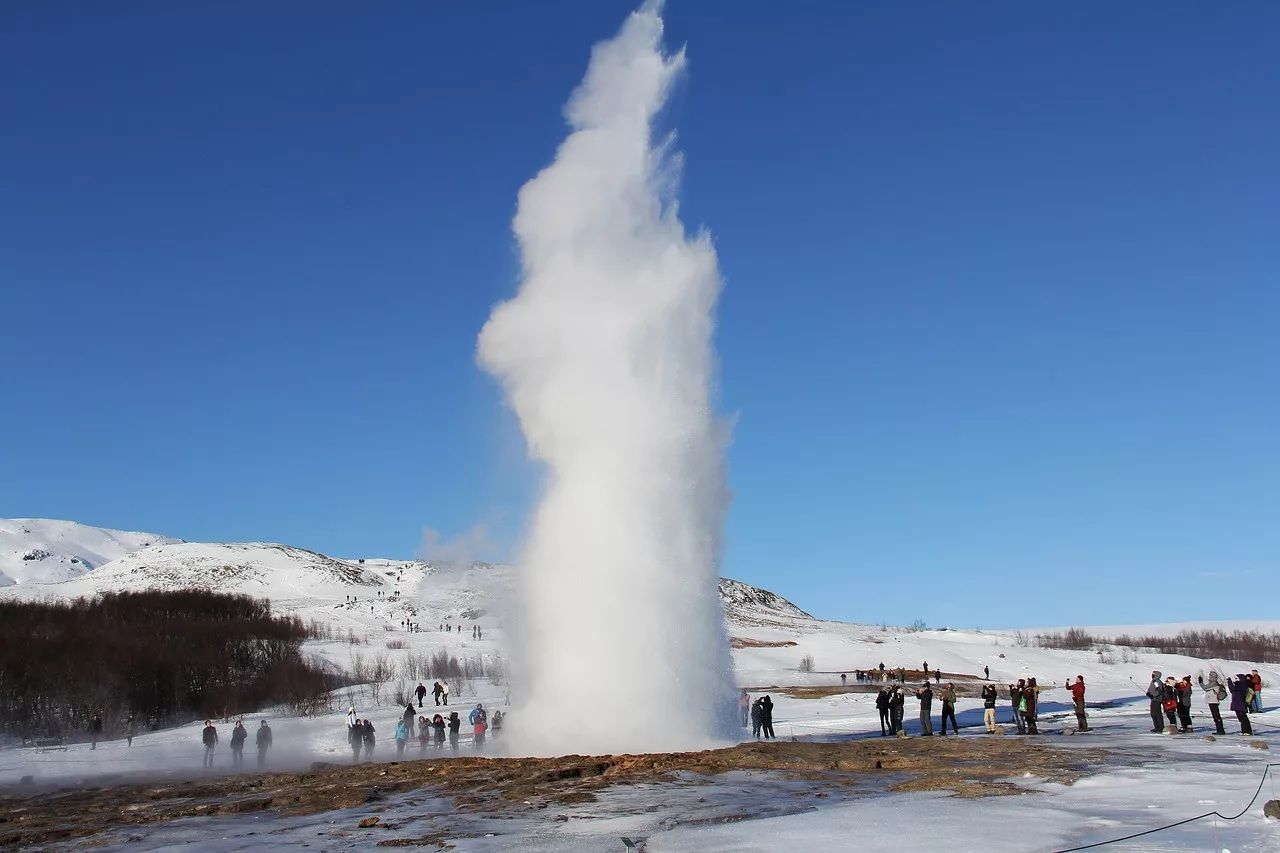 Un viaggio di gruppo WeRoad ammira un grande geyser che erutta in un paesaggio innevato sotto un cielo azzurro e limpido.