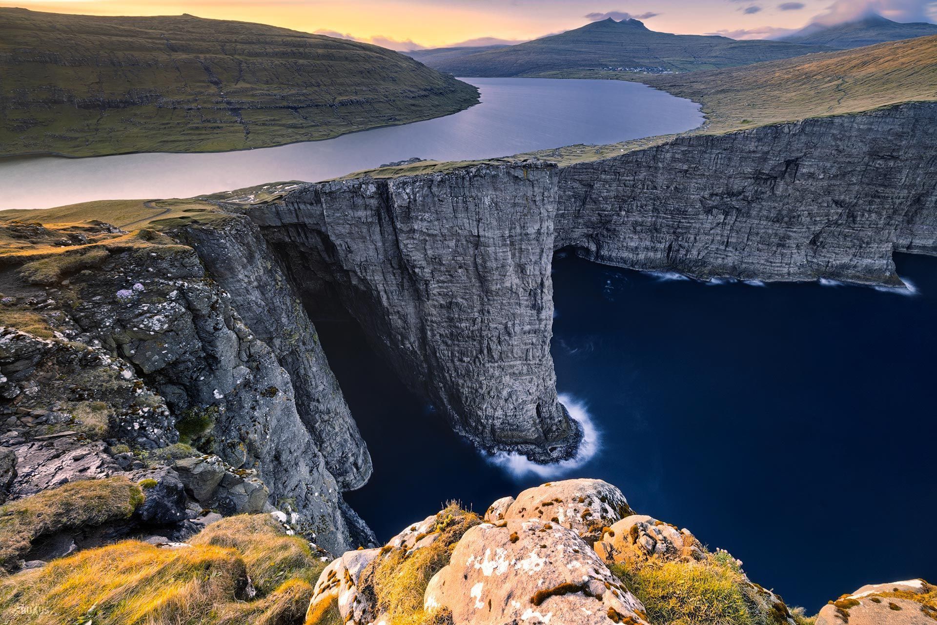 Un grande lago si trova sul bordo di un'alta scogliera, affacciato sul profondo oceano blu, con verdi colline ondulate in lontananza.