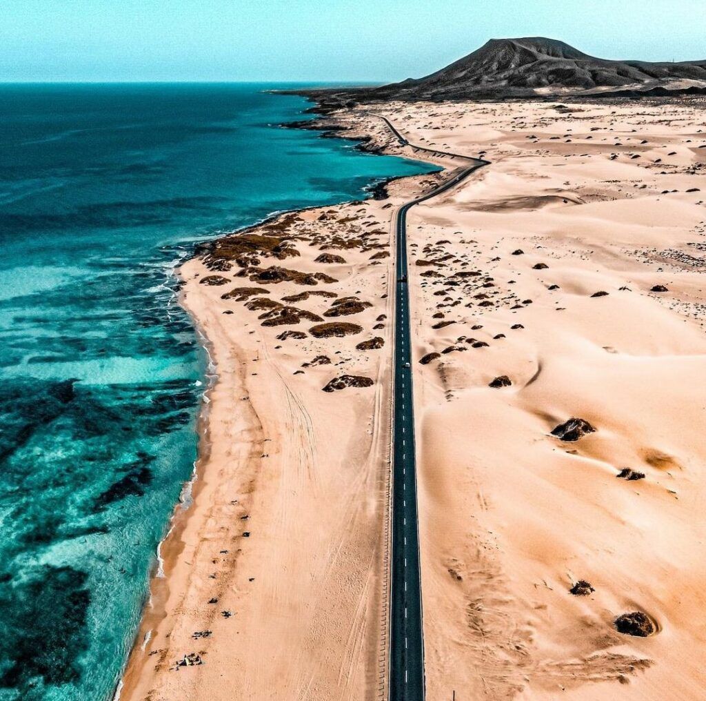 Una vista aerea di una strada asfaltata che corre tra un vasto deserto con una montagna e una costa oceanica turchese.