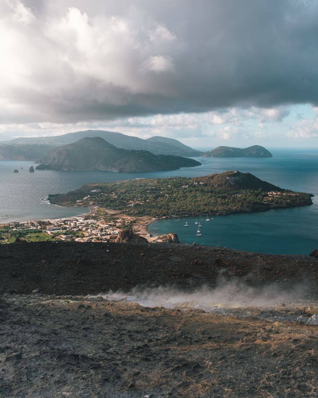 Una vista da un pendio vulcanico fumante che domina una città costiera su un'isola lussureggiante, circondata dal mare e da altre isole.