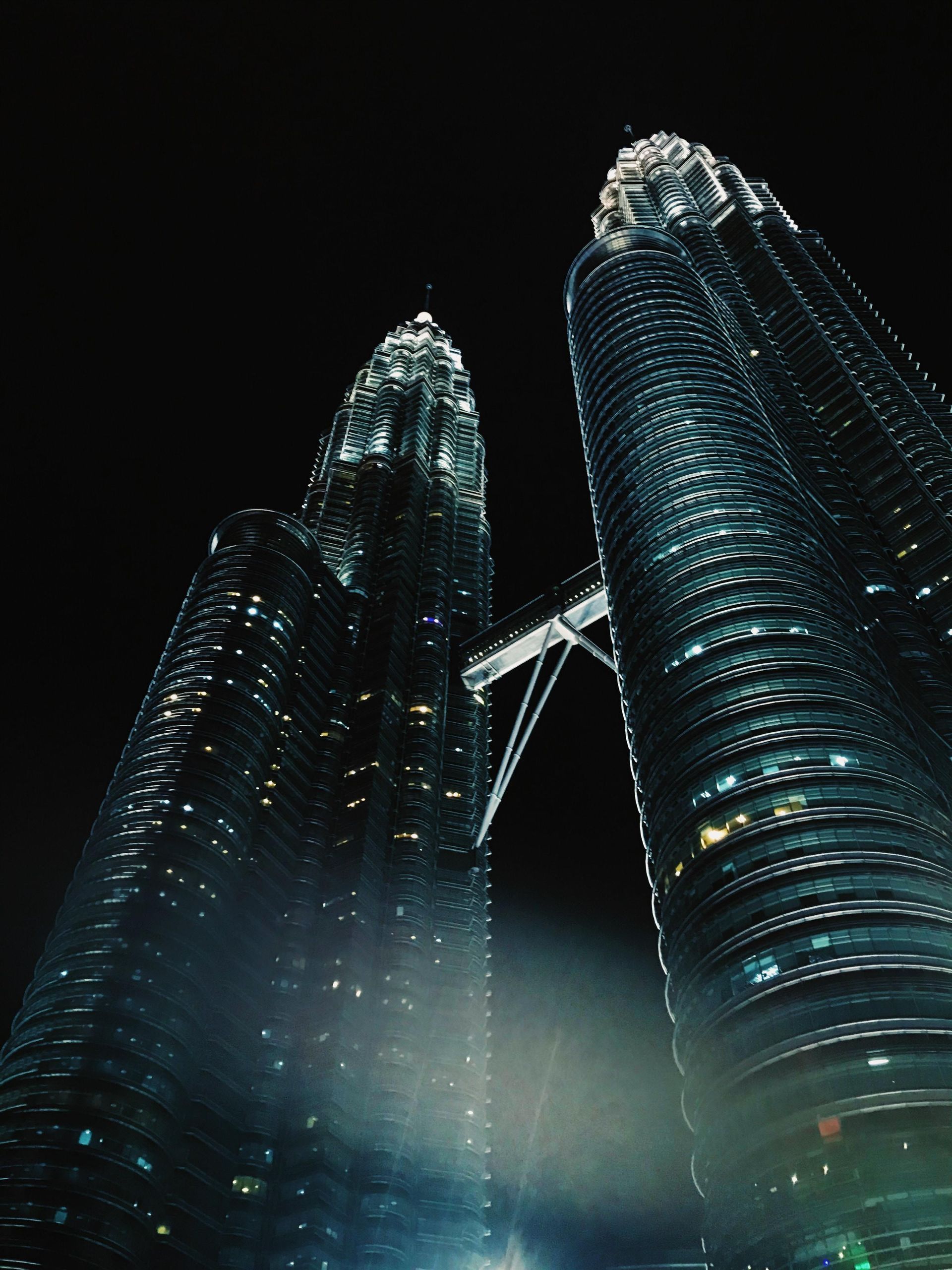Una vista en ángulo bajo de dos rascacielos modernos con un puente aéreo que los conecta, iluminados contra un cielo nocturno oscuro.