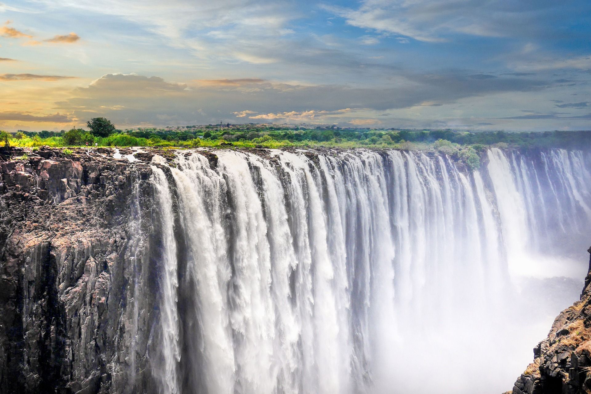 A massive waterfall rushes over a wide, rocky cliff edge, with green vegetation visible in the background under a blue and cloudy sky.