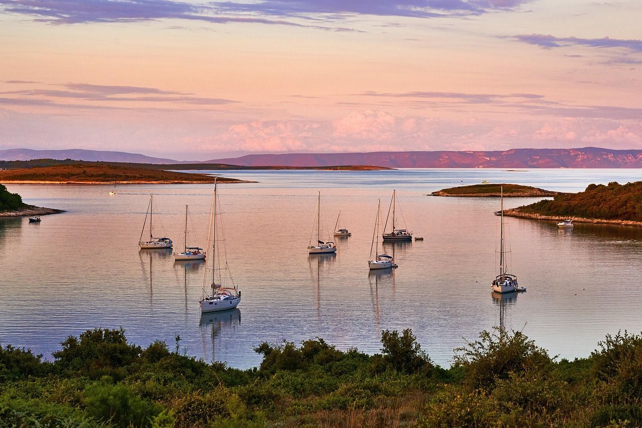 Several sailboats are anchored in a tranquil bay surrounded by islands under a pastel-colored sunset sky.