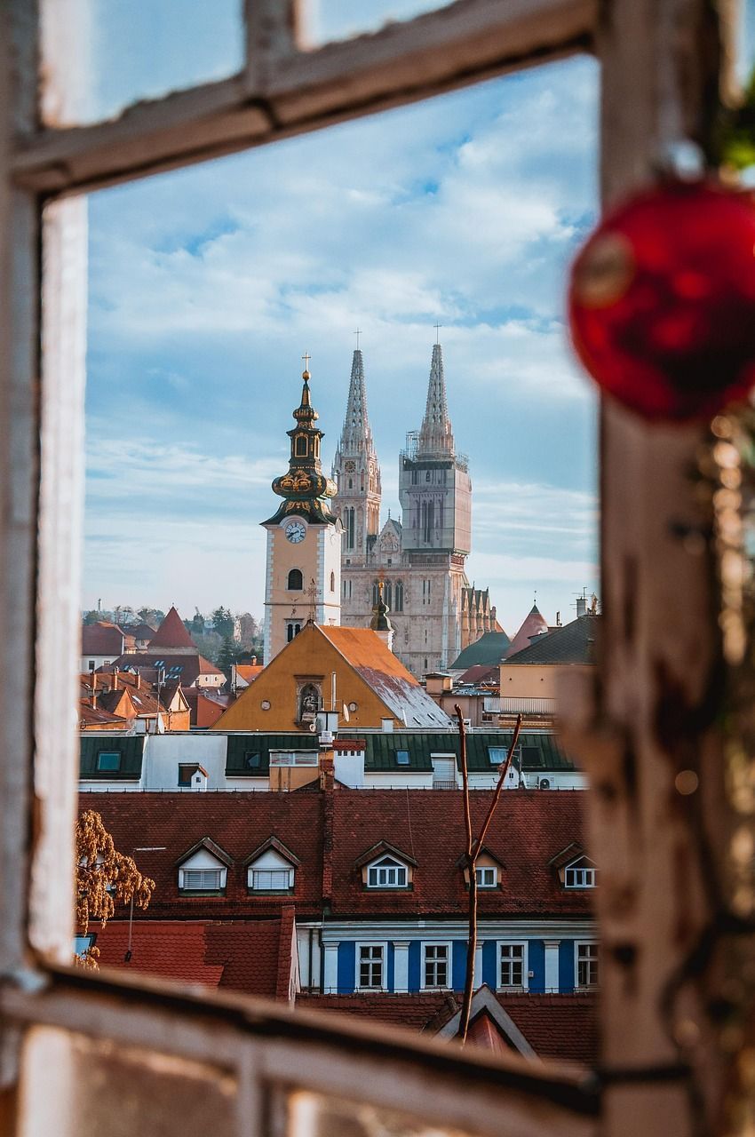 A cityscape featuring a cathedral and clock tower, viewed through a rustic window frame with a red ornament hanging in the foreground.