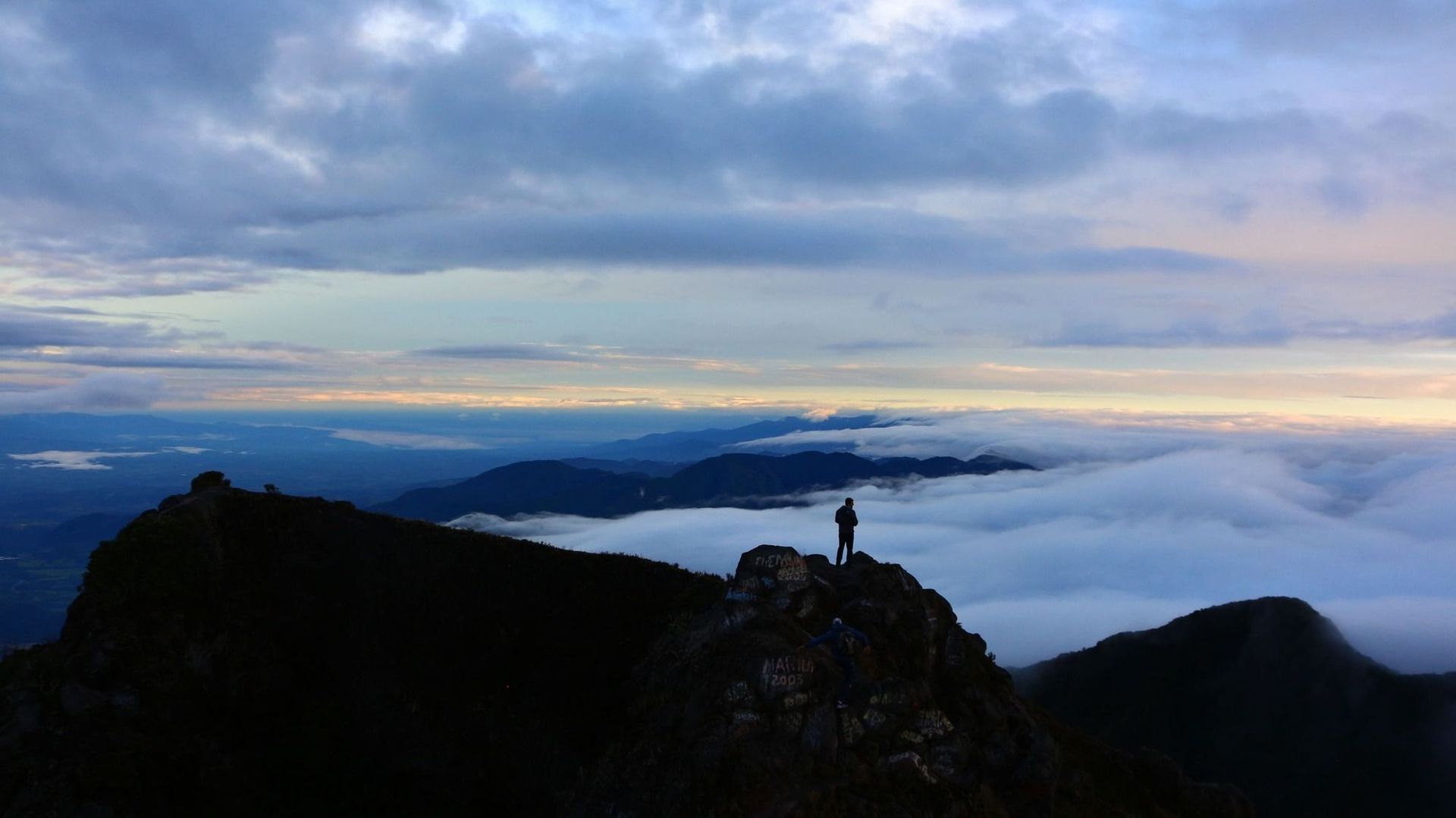 Una persona si staglia in silhouette su una vetta rocciosa di montagna, ammirando un vasto mare di nuvole e montagne lontane.