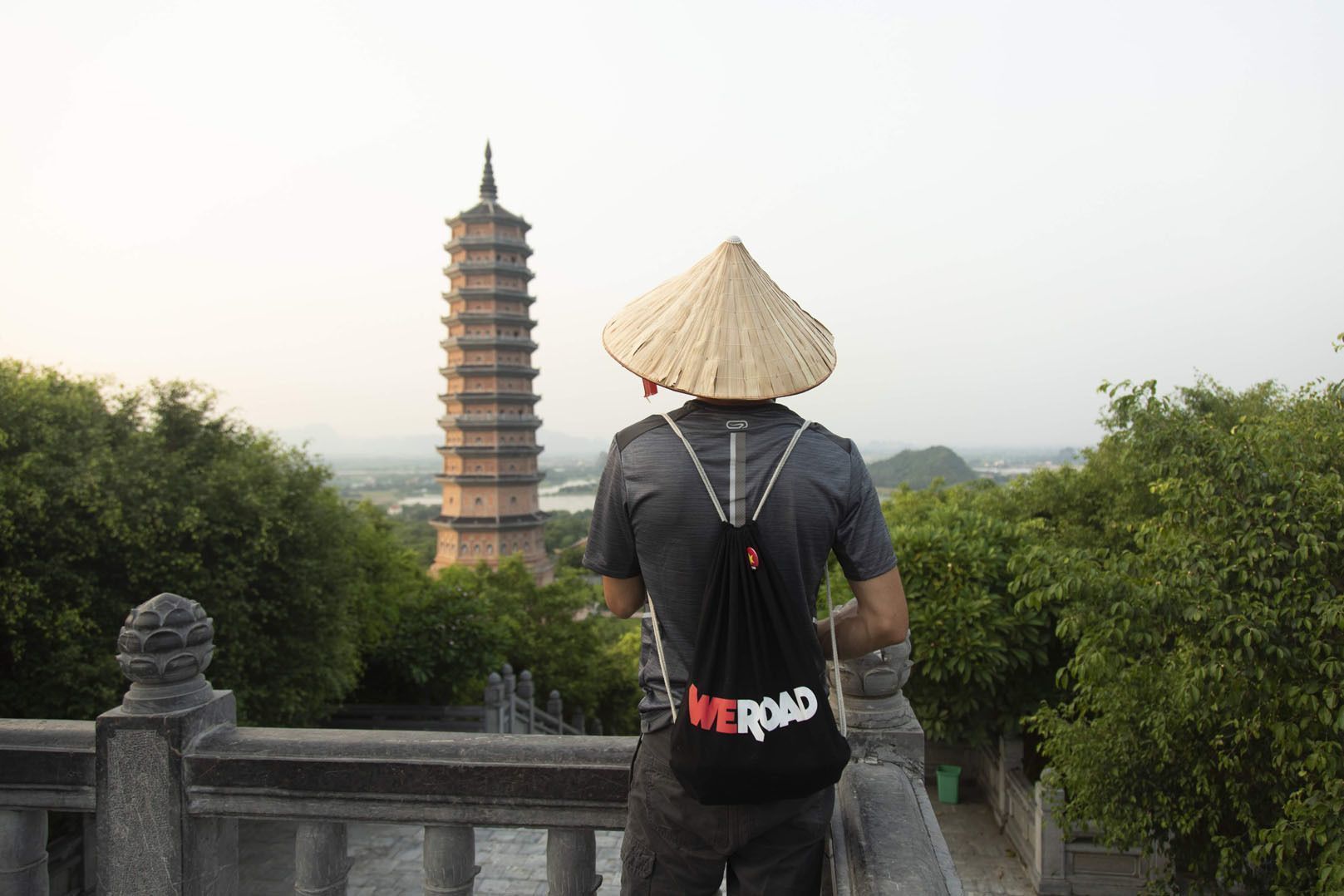 Un hombre de espaldas, con sombrero cónico y una mochila WeRoad, de pie en un balcón de piedra con vistas a una alta pagoda.