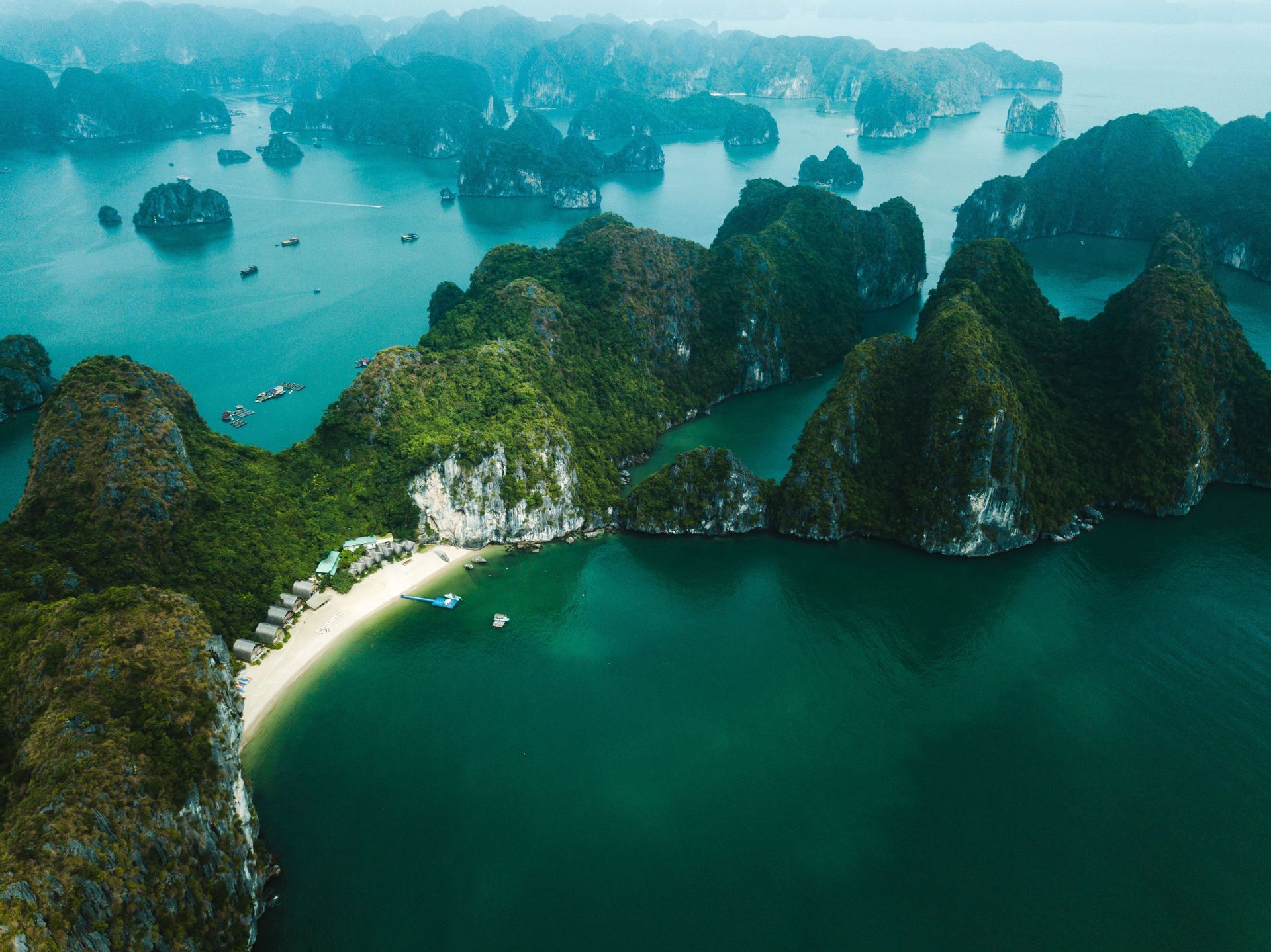 Aerial view of numerous green-topped limestone islands rising from turquoise water, with a secluded white sand beach and bungalows.