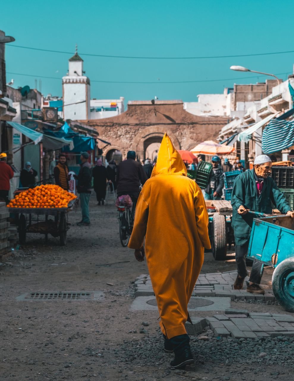 A person wearing a yellow hooded robe walks down a busy market street with a cart of oranges and a minaret in the distance.