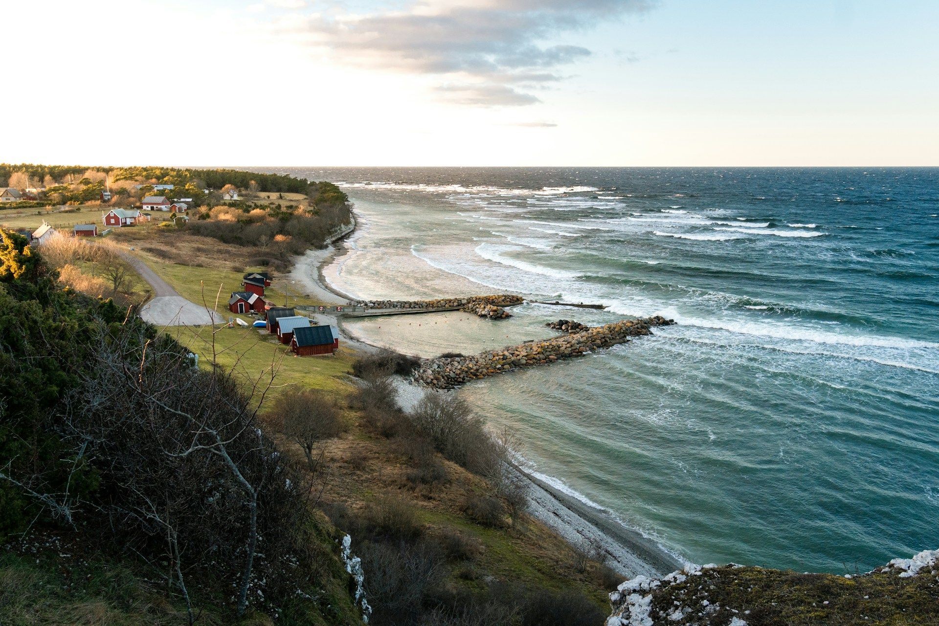 Blick von oben auf ein Küstendorf mit roten Häusern an einem grasbewachsenen Ufer, neben einem unruhigen Meer mit weißen Wellen.