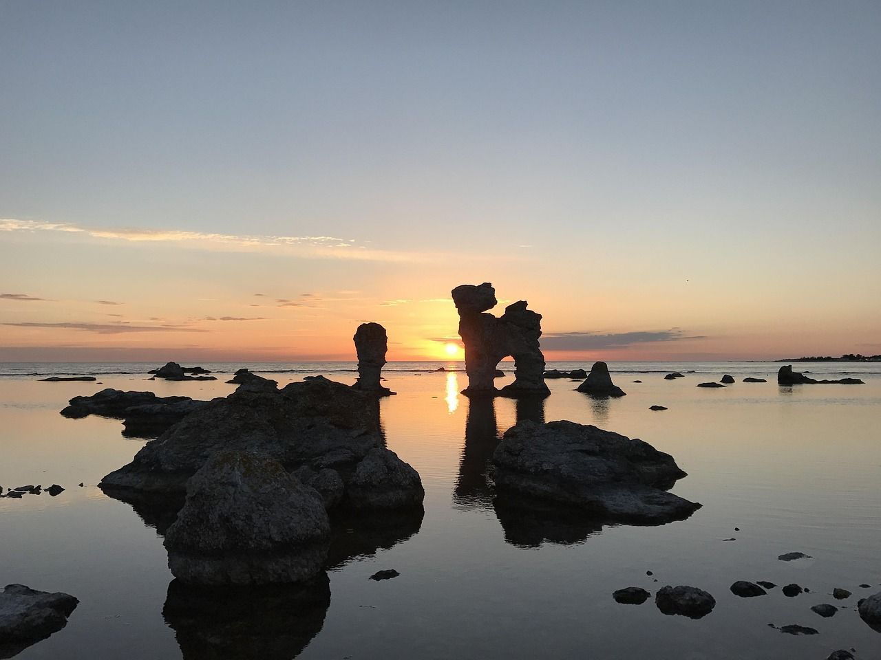 Silhouetten großer Felsformationen stehen im ruhigen Wasser, während die Sonne am Horizont untergeht.