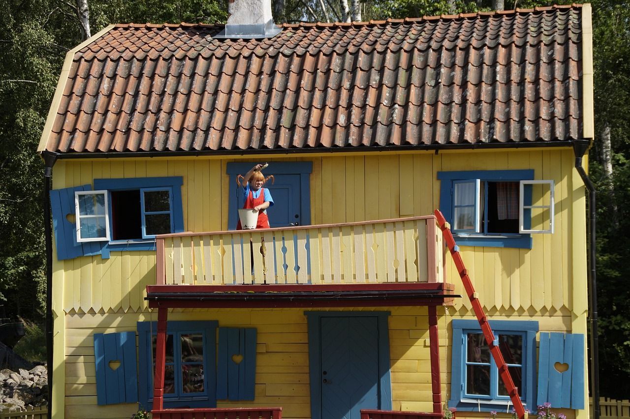 Eine Schaufensterpuppe von Pippi Langstrumpf steht auf dem Balkon eines gelben Hauses und hält einen weißen Eimer und einen Pinsel.