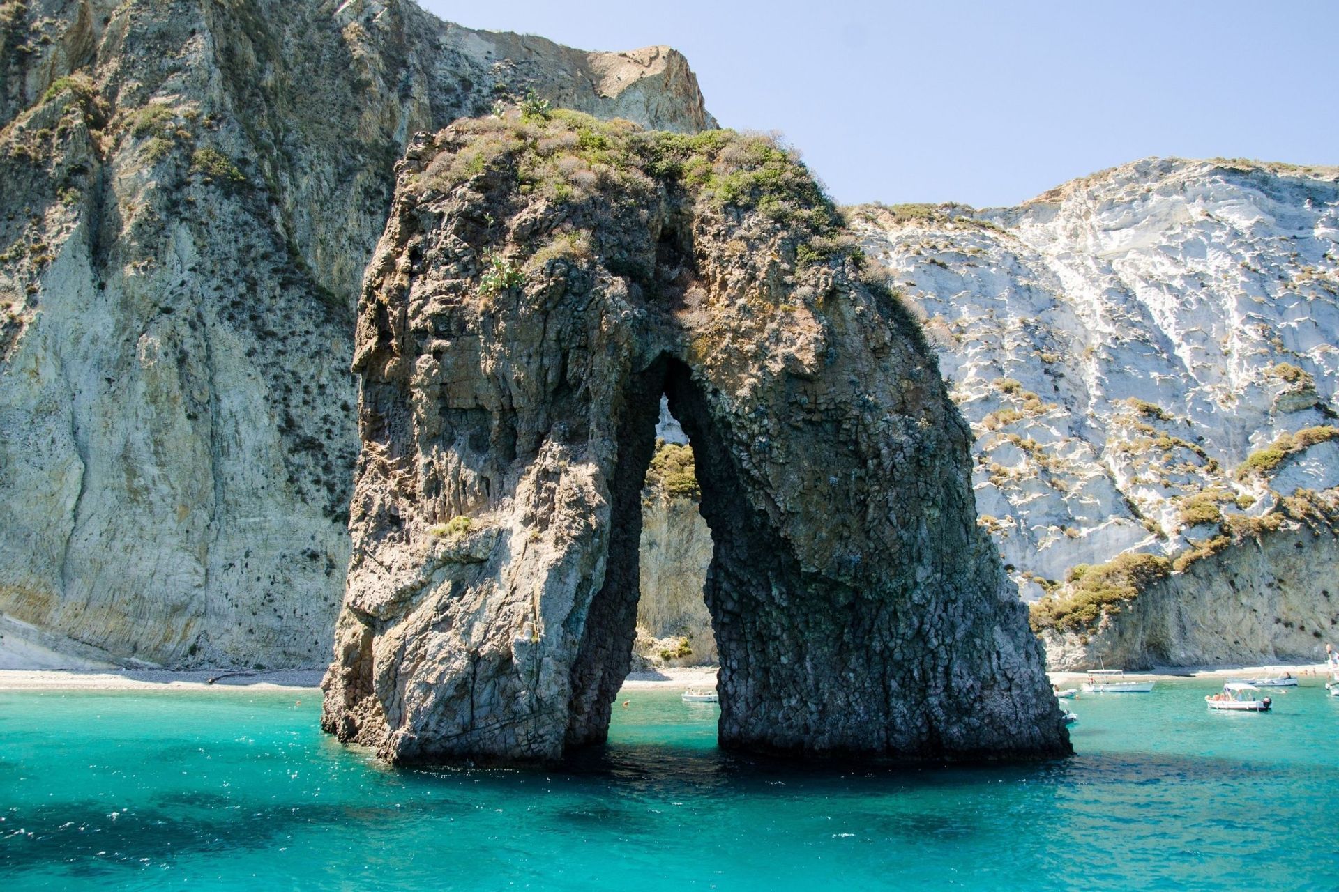 Un grande arco naturale di roccia si erge in acque turchesi, con una spiaggia e scogliere bianche sullo sfondo.