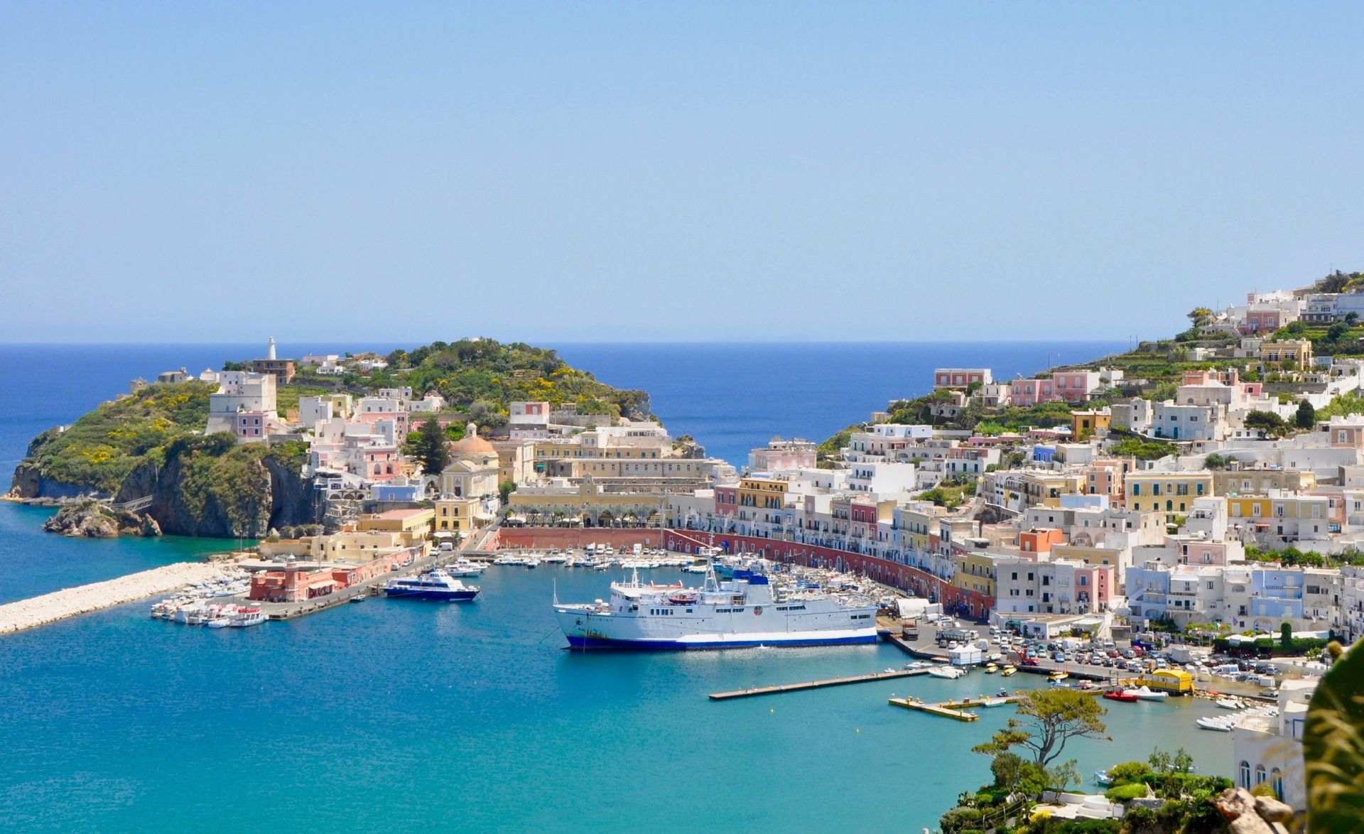 Una vista panoramica di una città di mare con edifici colorati sulle colline che si affacciano su un porto blu brillante con un grande traghetto.