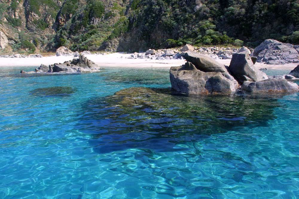 Acqua turchese limpida increspa grandi rocce davanti a una spiaggia isolata di sabbia bianca e una collina verdeggiante.
