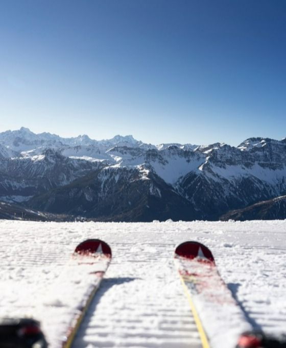 Una visuale in prima persona dalle punte degli sci su un pendio innevato, di fronte a un'ampia catena di montagne innevate sotto un cielo sereno.