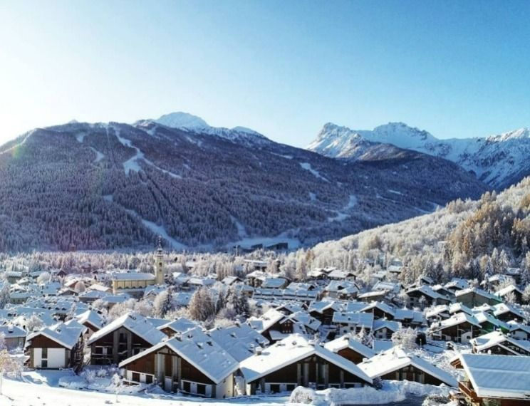 Una vista grandangolare di un villaggio alpino innevato con chalet incastonati ai piedi di montagne boscose in una giornata di sole.