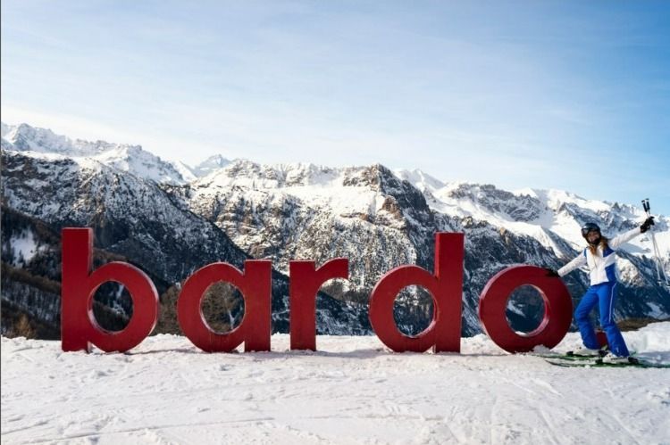 Una donna in tenuta da sci posa sugli sci accanto a grandi lettere rosse che formano la parola 'bardo' in un paesaggio montano innevato.