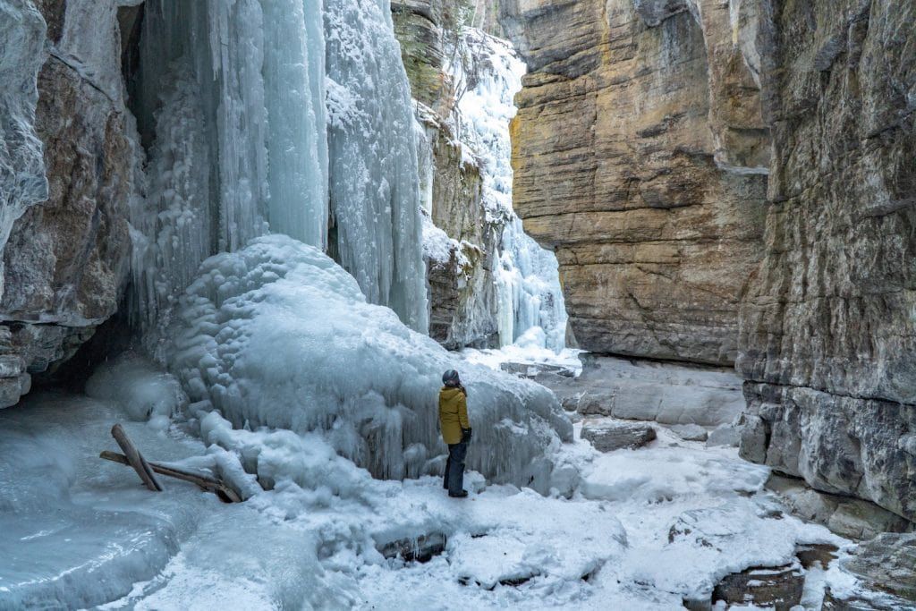 Una persona con una giacca invernale gialla si trova in un canyon innevato, guardando verso enormi cascate ghiacciate aggrappate alle pareti rocciose.