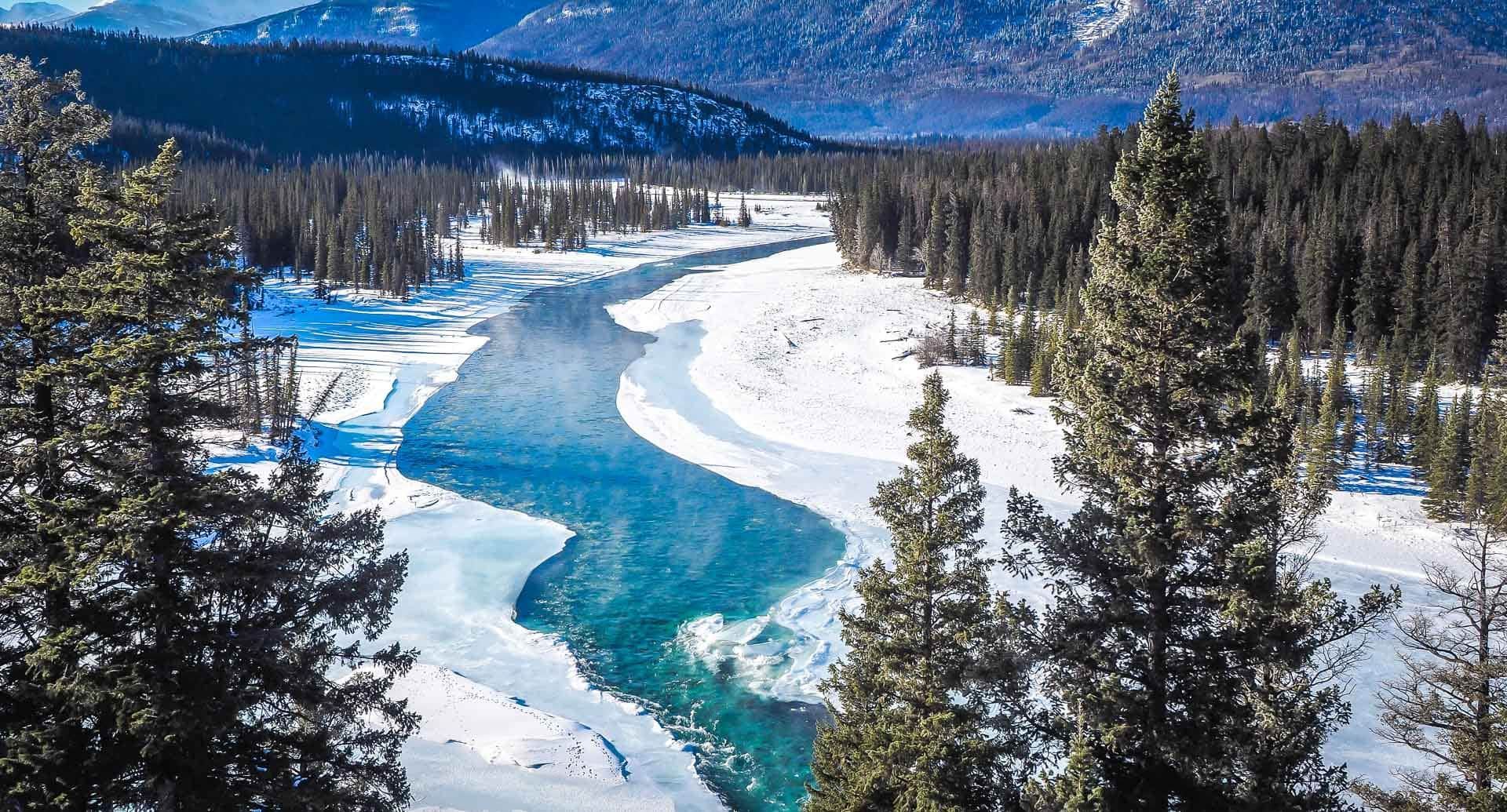 Un fiume azzurro brillante si snoda attraverso una valle innevata circondata da pini e montagne distanti.
