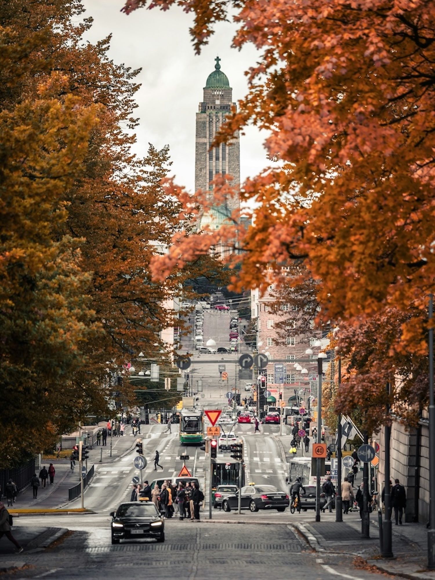 Una lunga strada di città, incorniciata da alberi con vivaci foglie autunnali arancioni, che si estende verso un'alta torre di chiesa in pietra in lontananza.