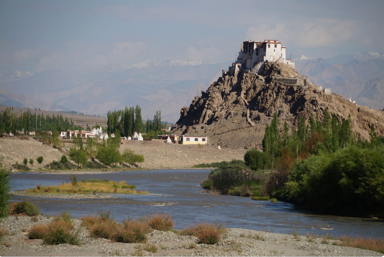 A white monastery sits on a rocky hill overlooking a river valley, with distant hazy mountains in the background.