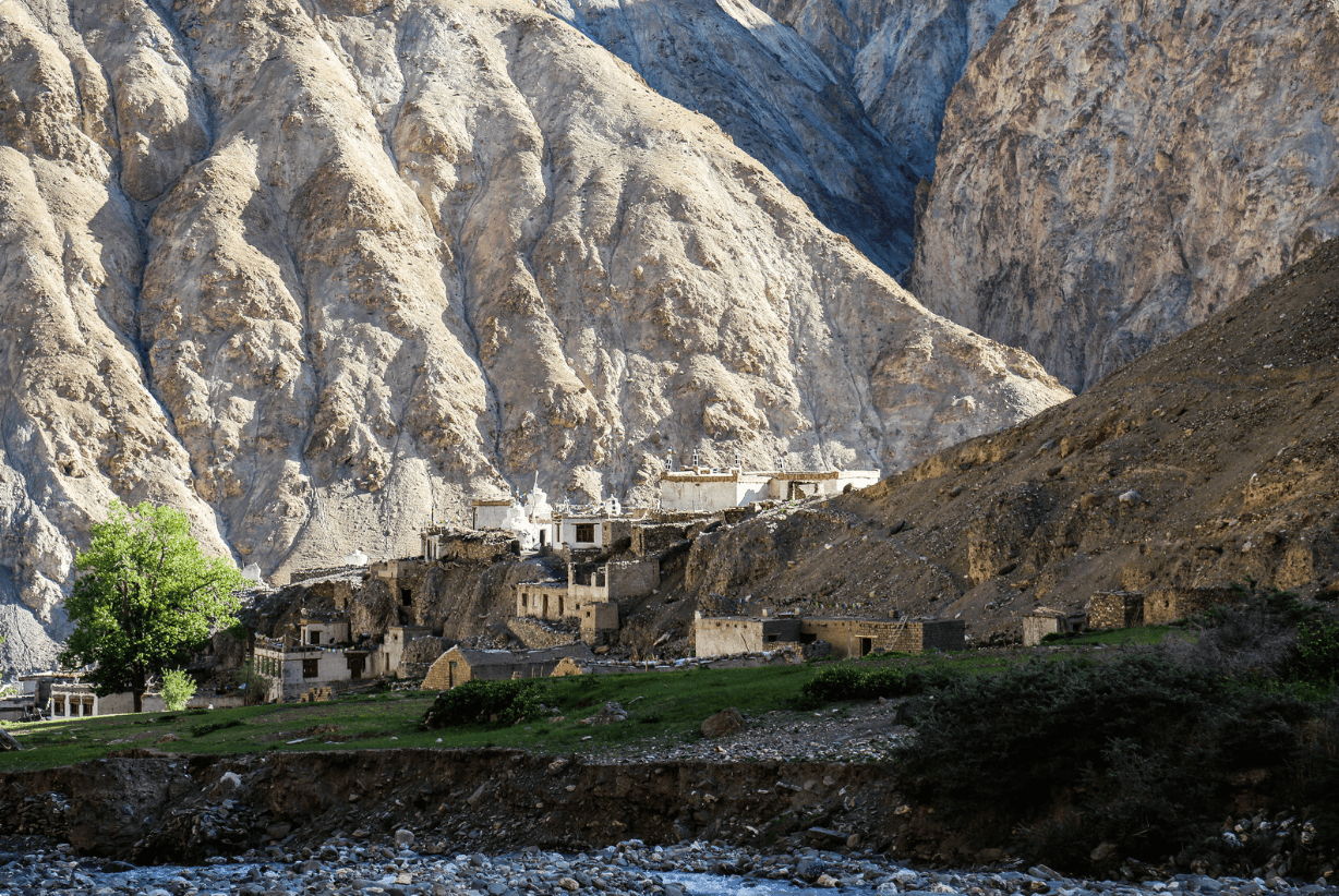 A small stone village with white-topped buildings is nestled into a grassy hillside at the base of a large, rugged mountain.