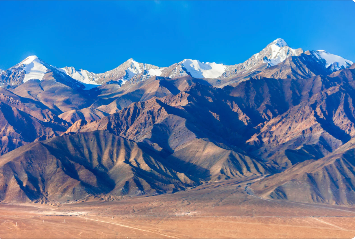 A rugged mountain range with snow-capped peaks stands against a clear blue sky, with a vast desert landscape in the foreground.