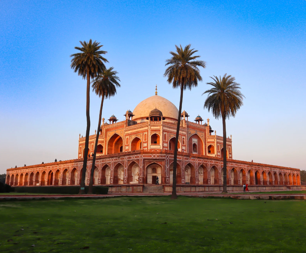 A red and white stone mausoleum with a central dome stands behind a green lawn, with four tall palm trees in front under a clear sky.