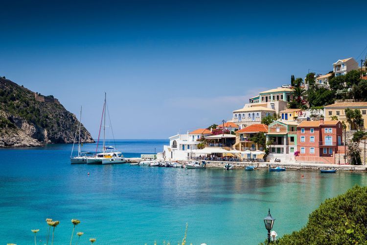 Case colorate di un villaggio marinaro costruito su una collina che si affaccia su una baia con acqua turchese e barche ormeggiate.