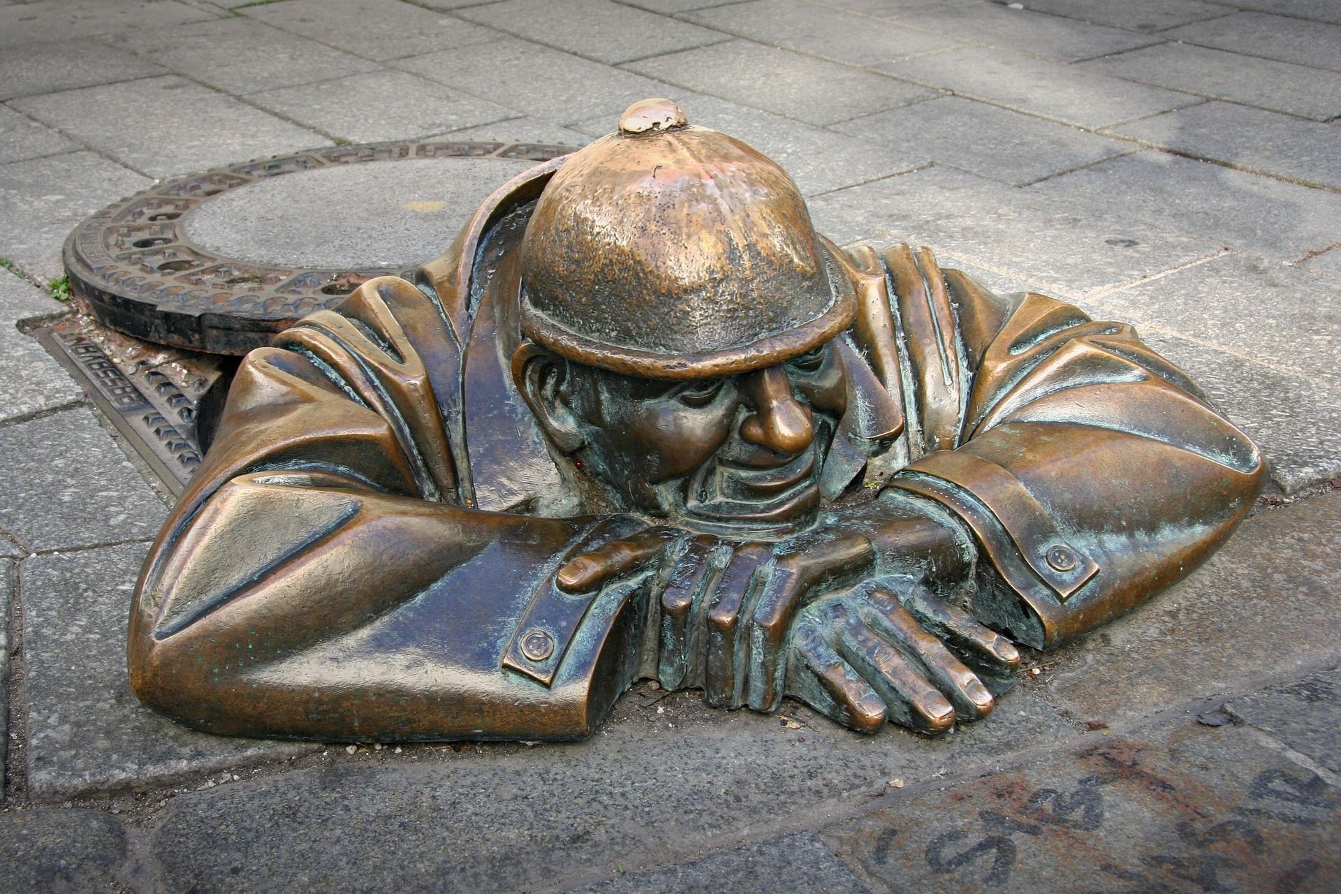 Una estatua de bronce de un hombre con sombrero emerge de una alcantarilla abierta en una acera urbana pavimentada, apoyando los brazos en el suelo.