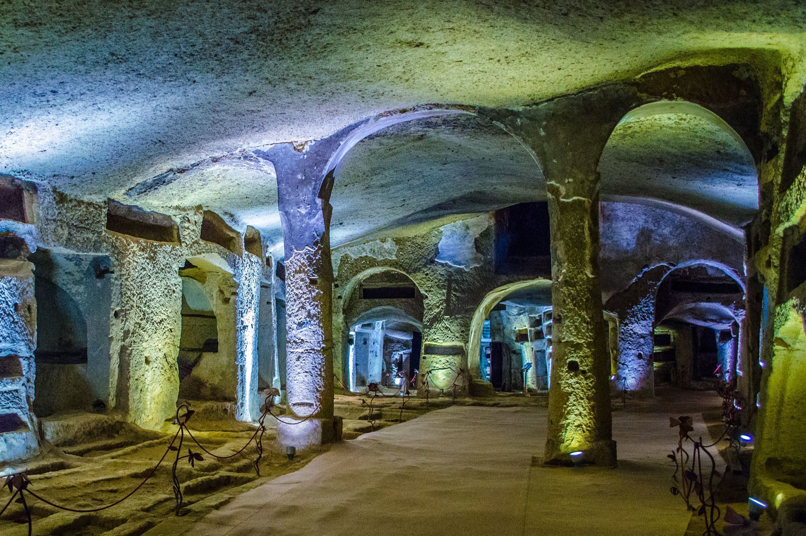 A large underground catacomb with rough-hewn stone pillars and arches, illuminated by blue and yellow lights.