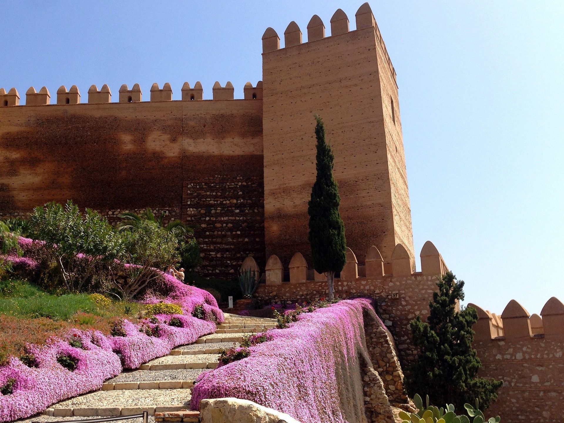 Una escalera de piedra bordeada por cascadas de flores moradas conduce a una gran fortaleza antigua con almenas contra un cielo azul claro.