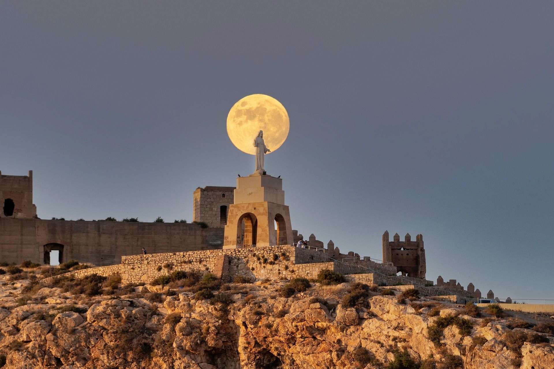 Una gran luna llena se alinea perfectamente detrás de una estatua sobre un pedestal en un castillo de piedra, sobre una colina rocosa al anochecer.