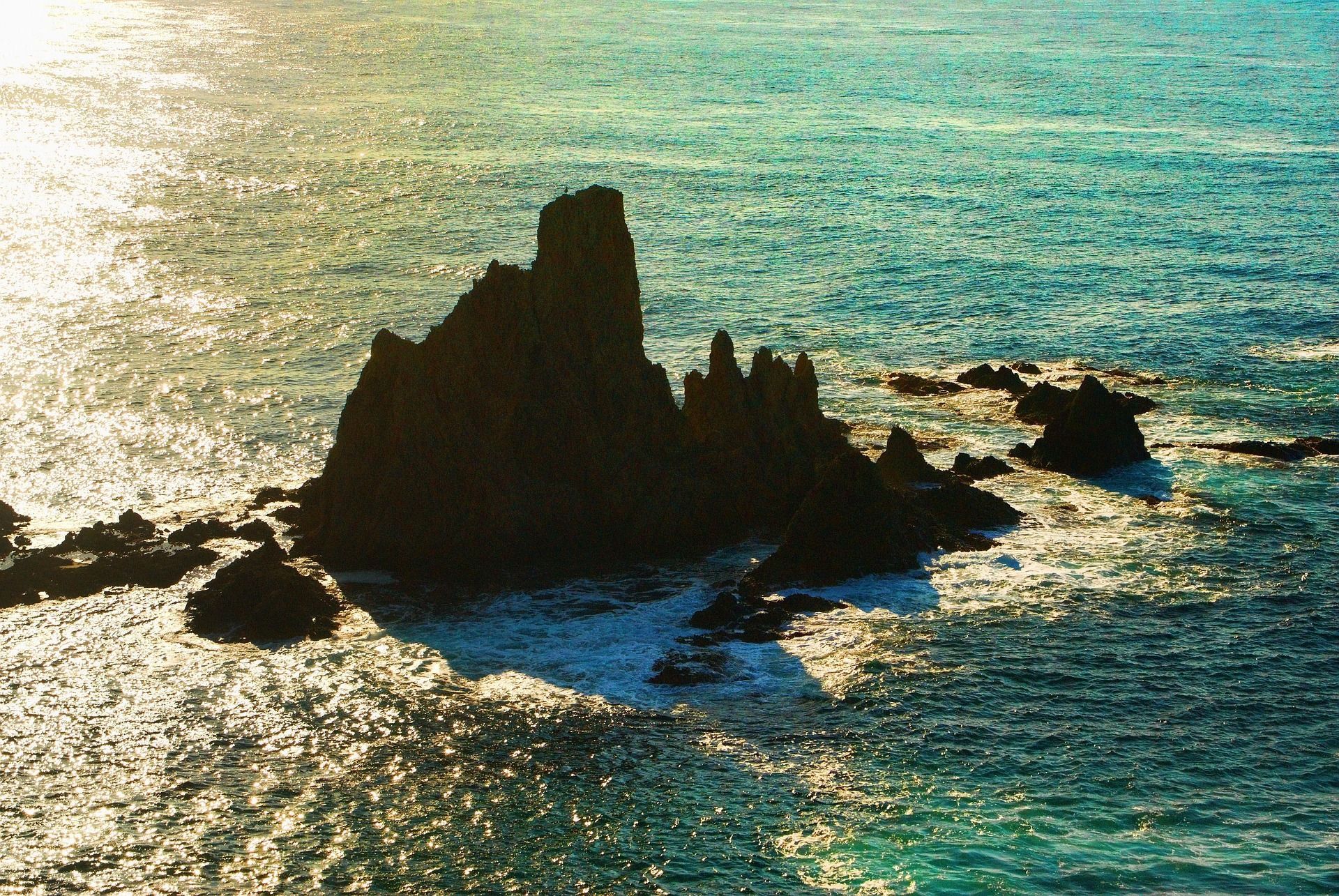 Un cúmulo de rocas oscuras y dentadas se eleva del mar turquesa, rodeado de agua brillante por el sol y espuma blanca.