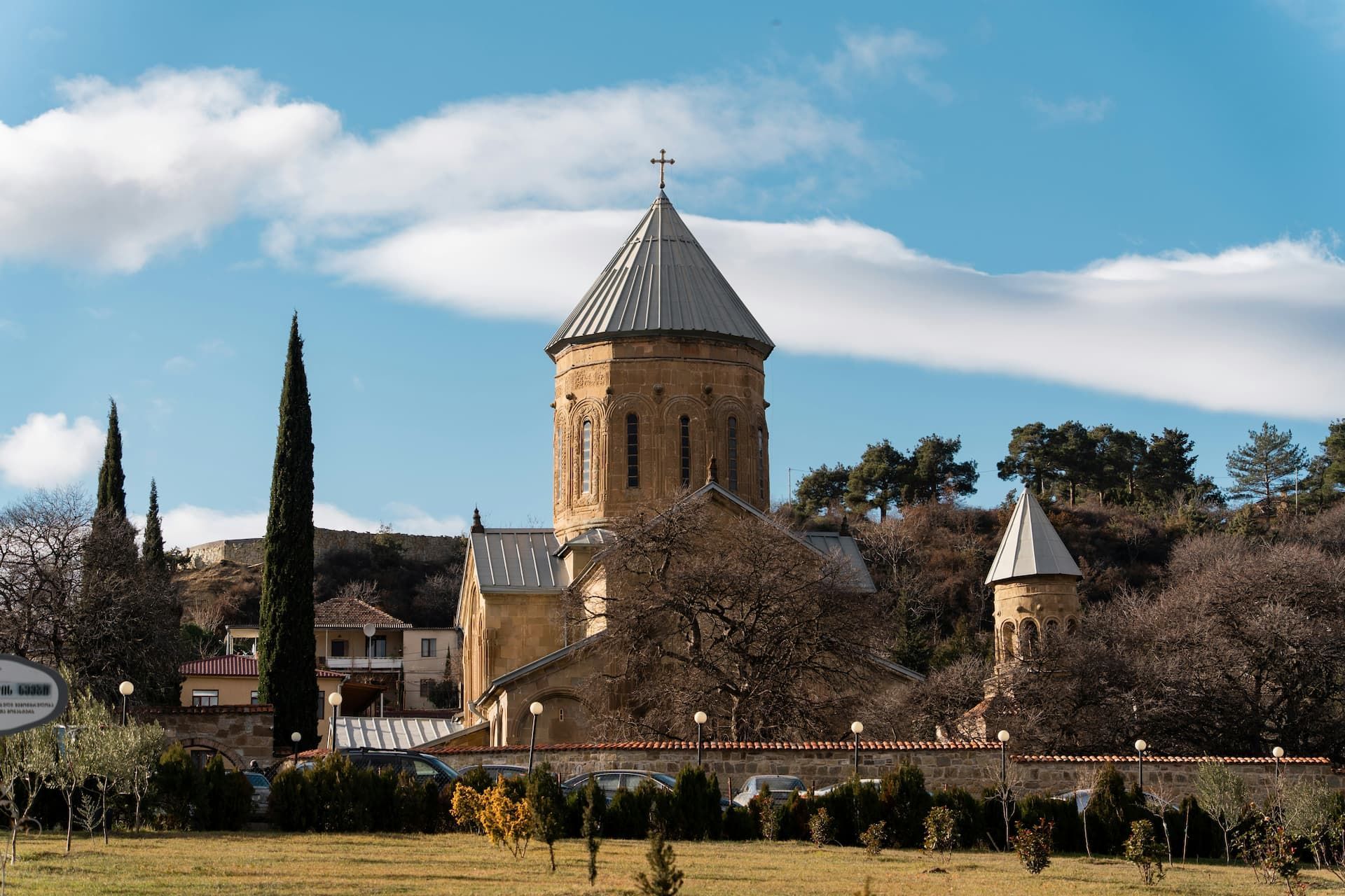 Un monastère en pierre avec un toit conique se dresse sur une pelouse herbeuse, entouré d'arbres, sous un ciel partiellement nuageux.