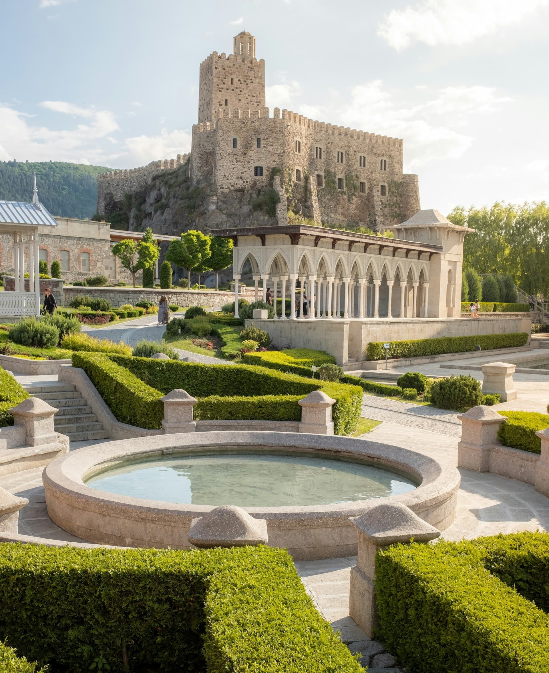 Un château de pierre sur une colline rocheuse surplombe un jardin à la française avec une fontaine ronde, des haies taillées et un pavillon de pierre.