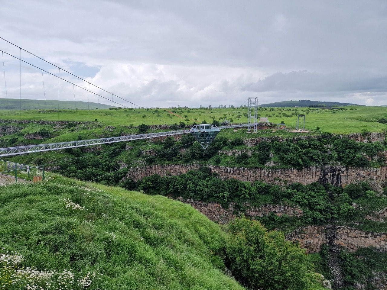 Un pont suspendu en verre avec une structure en forme de diamant au centre enjambe un canyon profond et verdoyant sous un ciel nuageux.