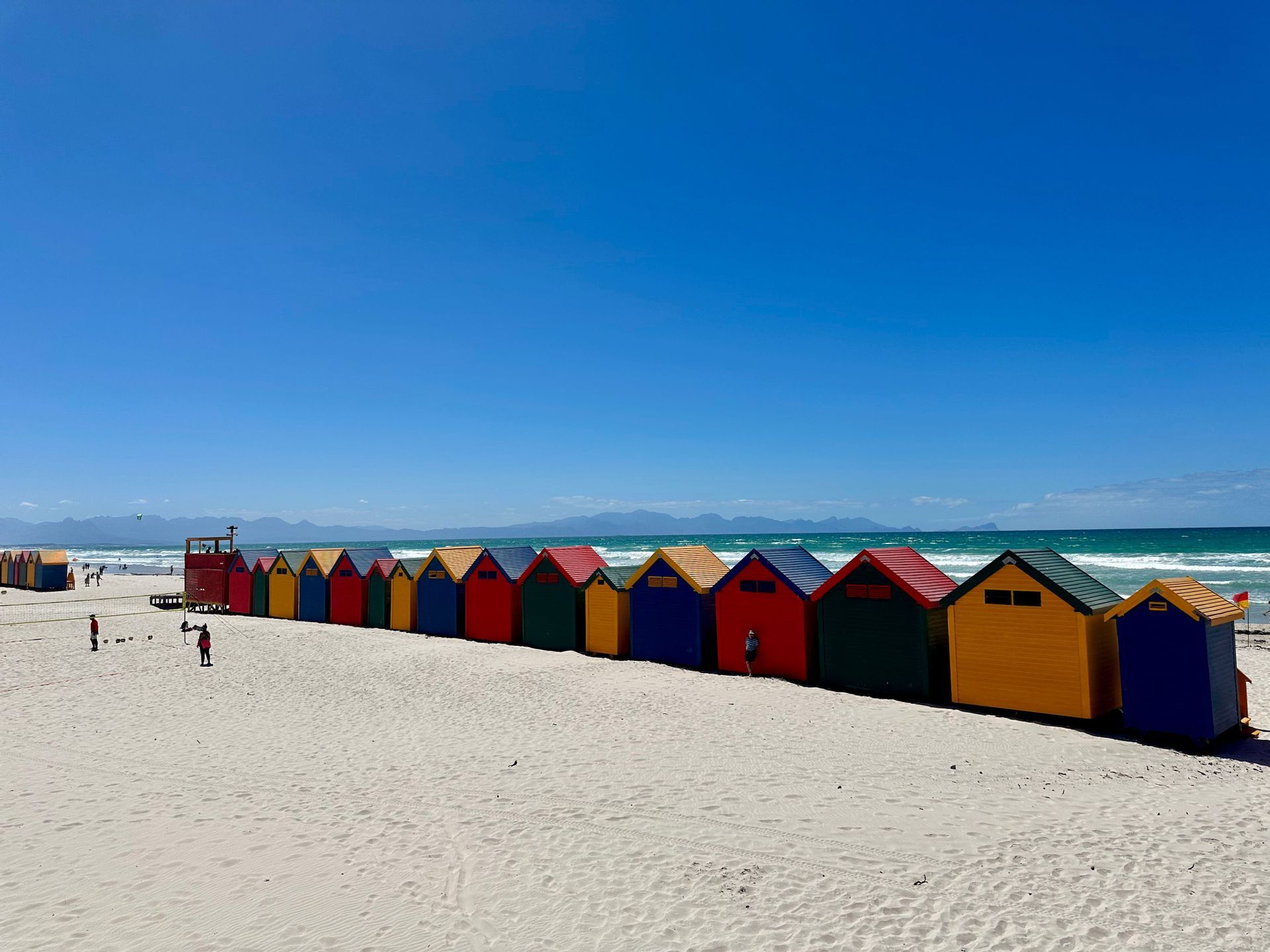Una lunga fila di colorate capanne di legno su una spiaggia di sabbia bianca con l'oceano sullo sfondo e un cielo sereno.