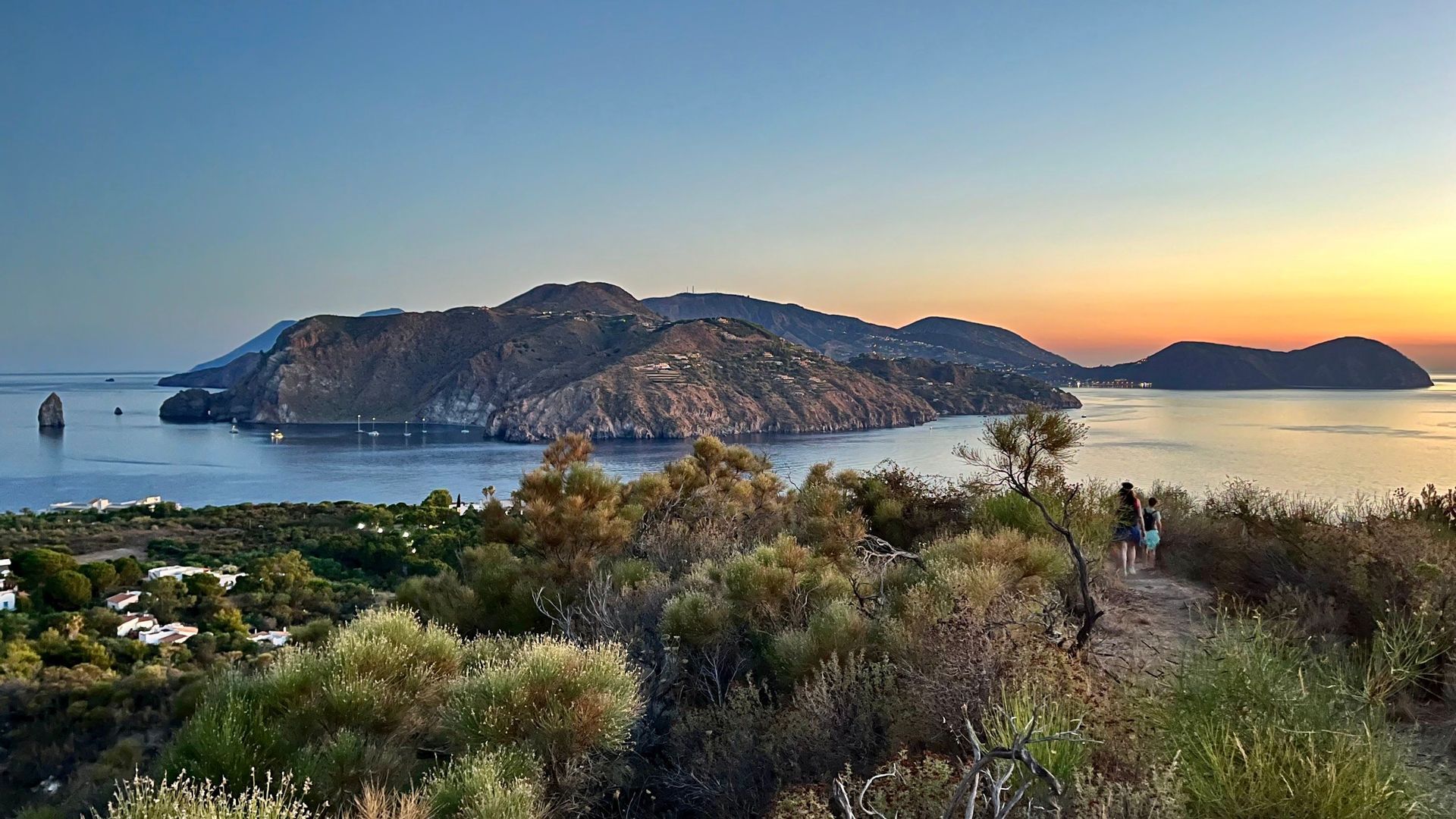 Due persone di un viaggio di gruppo WeRoad camminano su un sentiero collinare che domina una baia con isole vulcaniche al tramonto.