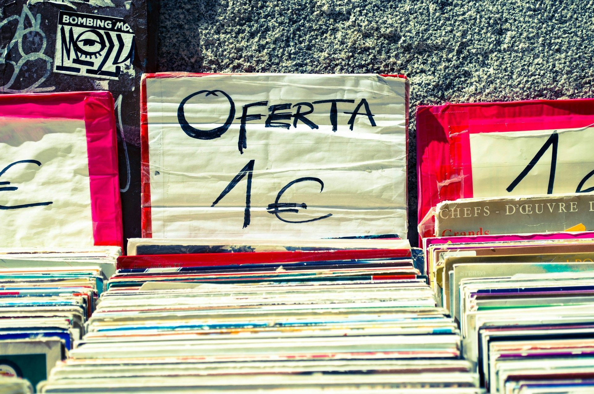 A handwritten sign reading 'Oferta 1€' sits atop stacks of secondhand vinyl records at an outdoor market stall.