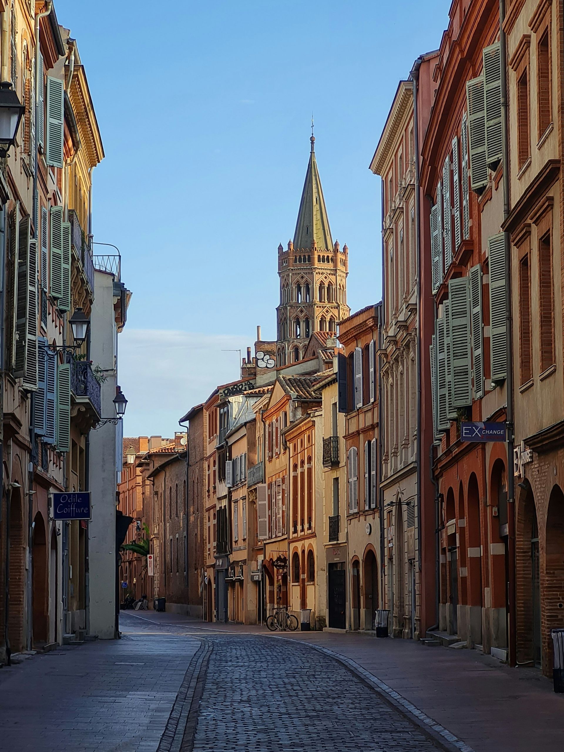 Une rue pavée vide serpente à travers de vieux bâtiments de style européen, menant à un haut clocher sous un ciel bleu.