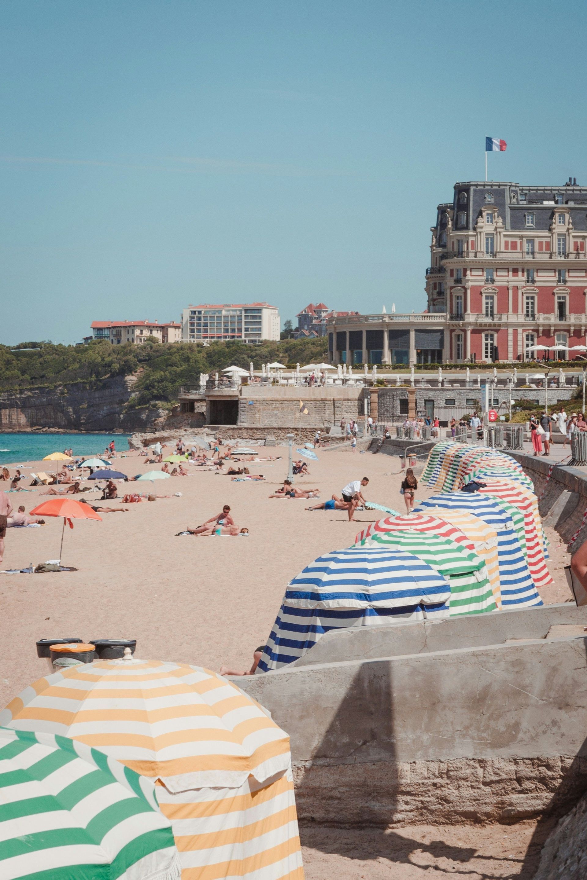 Des gens se prélassent au soleil sur une plage de sable remplie de parasols rayés colorés, devant un grand bâtiment arborant un drapeau français.
