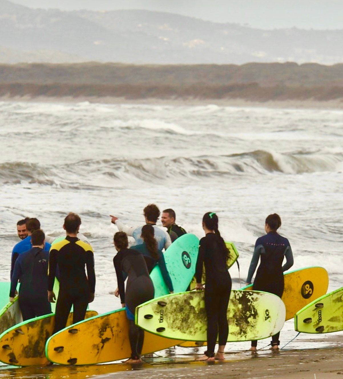 A WeRoad group trip wearing wetsuits stands on a beach with surfboards, looking at the ocean waves before a surf lesson.