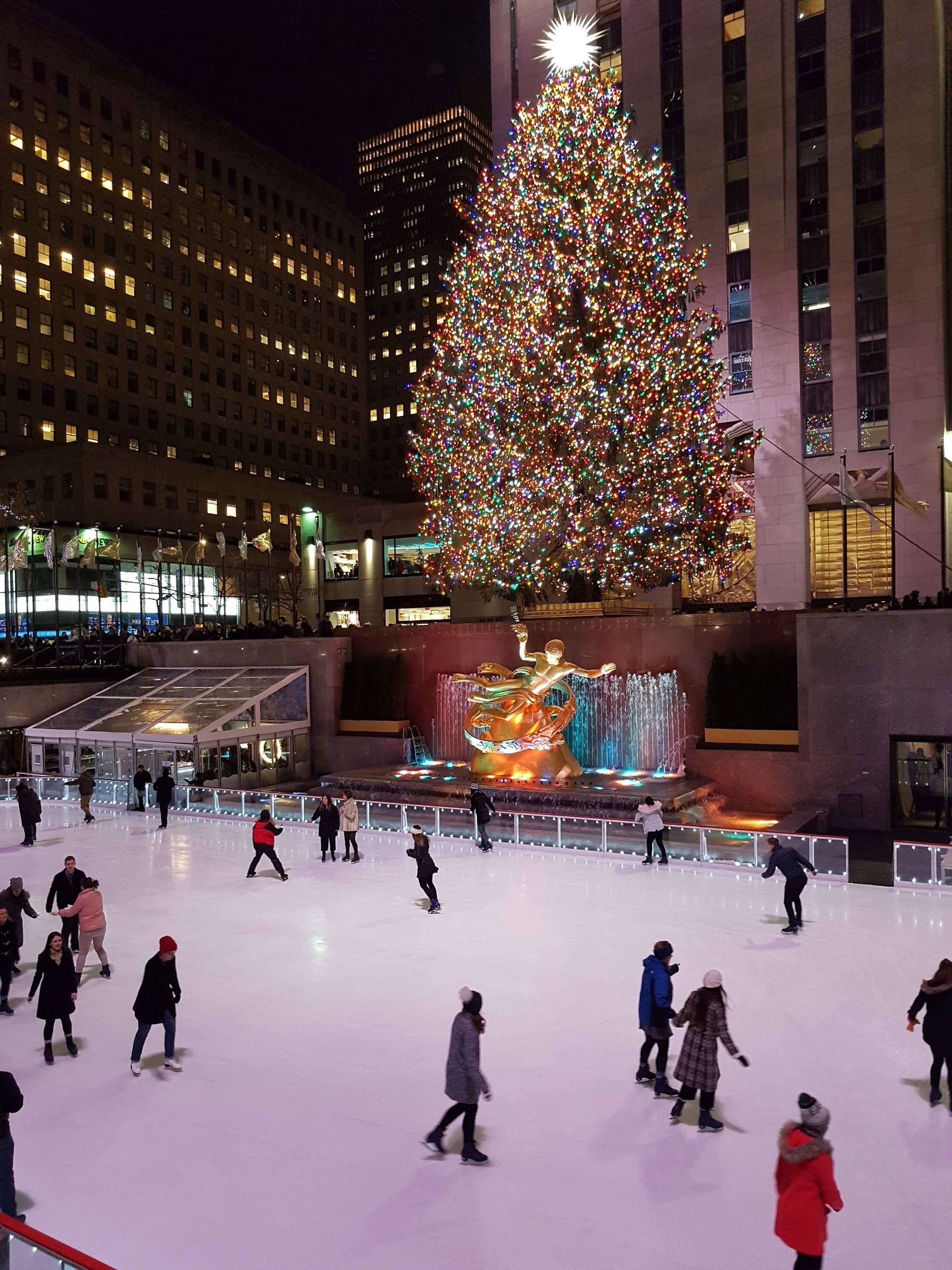 Menschen laufen nachts auf einer Open-Air-Eisbahn in einer Stadt, vor einem großen, hell erleuchteten Weihnachtsbaum und einer goldenen Statue.