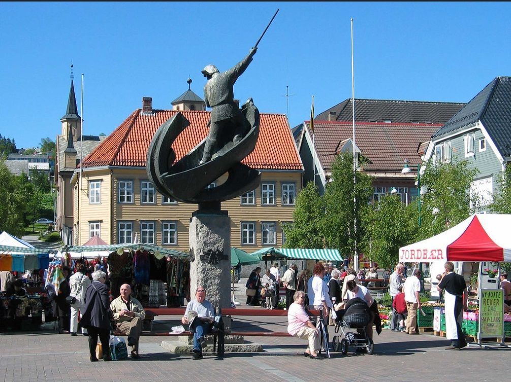 La gente se reúne en un mercado al aire libre en una plaza, centrado alrededor de una gran estatua de un hombre en un barco.