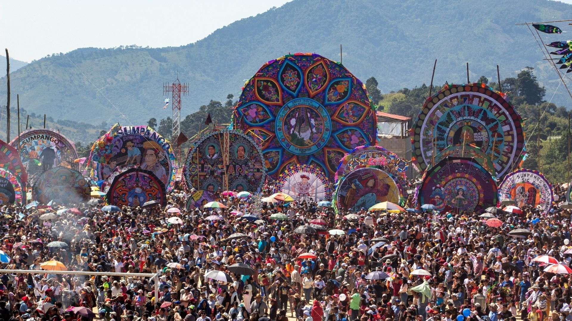 Eine riesige Menschenmenge versammelt sich auf einem Open-Air-Festival, um mehrere riesige, bunte, verzierte Drachen zu bestaunen, die vor einer Bergkette ausgestellt sind.