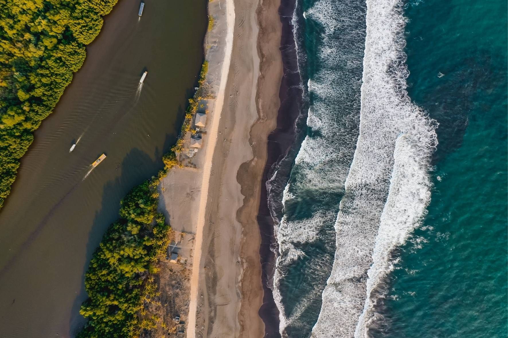 Eine Luftaufnahme einer Küstenlinie, die einen Fluss mit Booten zeigt, getrennt vom türkisfarbenen Meer durch einen schmalen Sandstrand.
