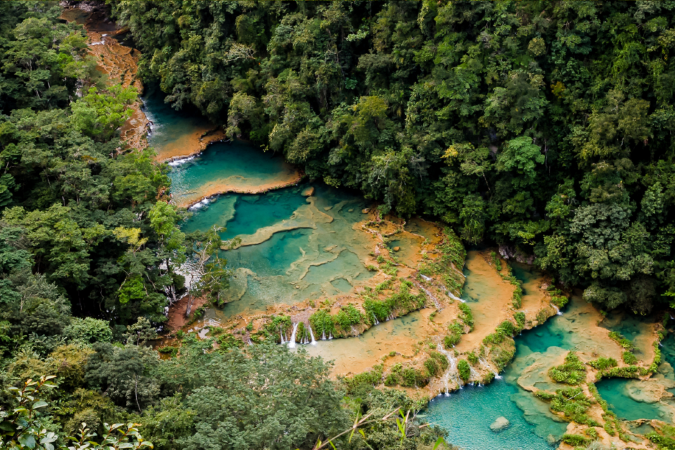 Luftaufnahme eines türkisfarbenen Flusses mit terrassierten Becken und kleinen Wasserfällen, der durch einen dichten grünen Dschungel fließt.