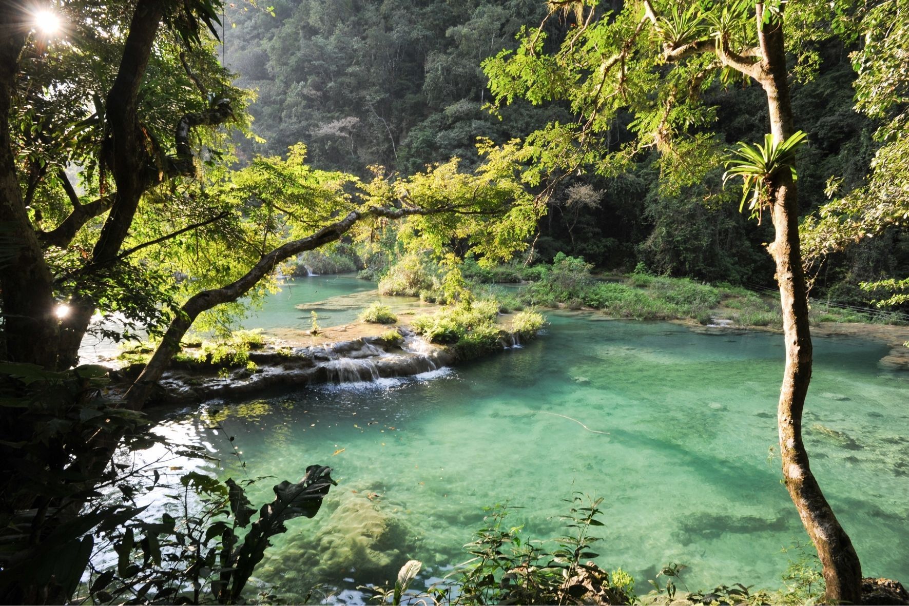 Die Sonne scheint durch üppige Dschungelbäume über kaskadierende Wasserfälle, die in einen klaren, türkisfarbenen Fluss münden.